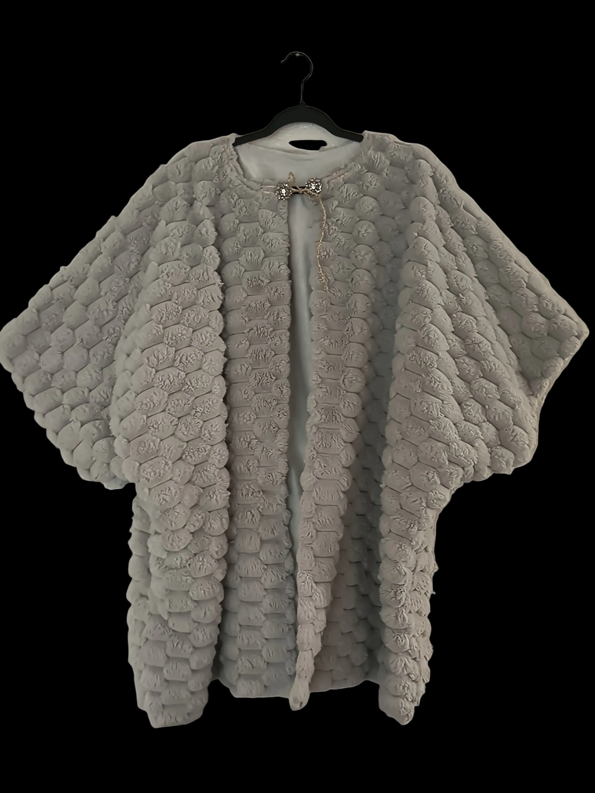 A light gray textured poncho-style jacket with a 3D rosette pattern, displayed on a black hanger against a solid black background.