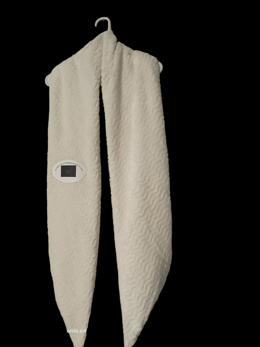 A white textured fleece neck warmer or scarf hanging from a metal hook on a textured beige wall. A small digital temperature strip is attached to the left side.
