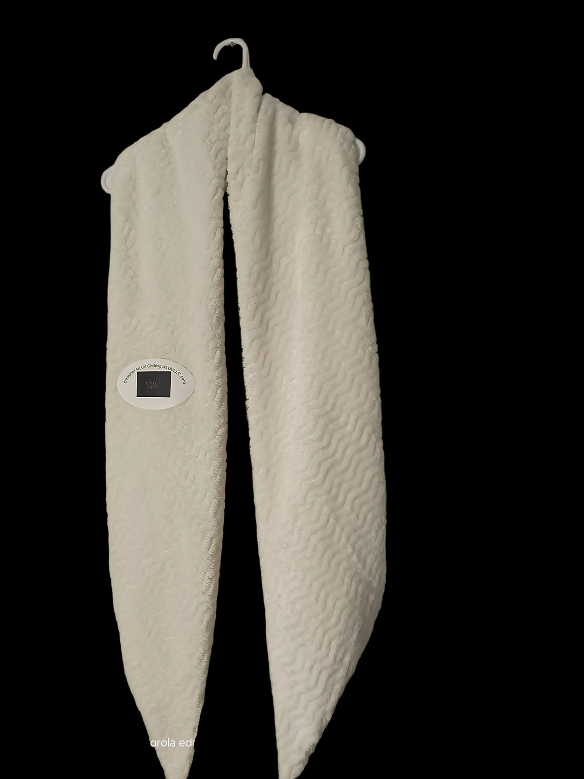 A white textured fleece neck warmer or scarf hanging from a metal hook on a textured beige wall. A small digital temperature strip is attached to the left side.